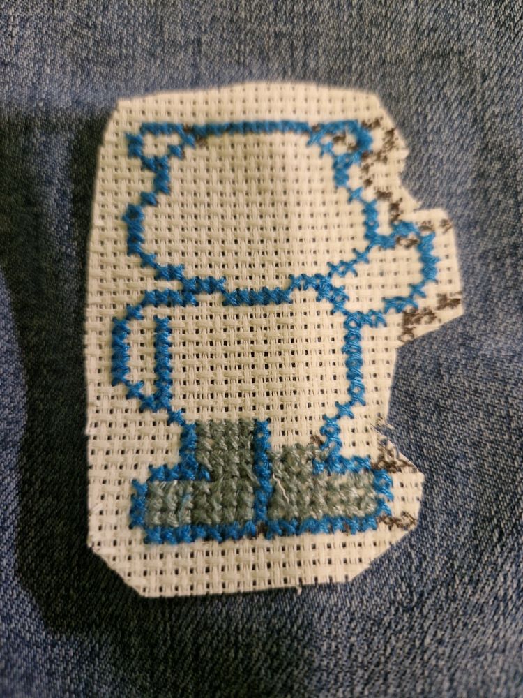 Omrisset av en bamse brodert med blå korssting på Aida på jeans. Føttene er fylt inn med en sjøgrønn farge. 