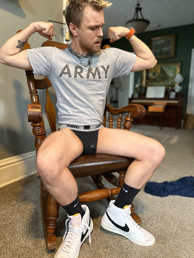 Troy sits on a wooden rocking chair, wearing an ARMY PT shirt, black BIKE jockstrap, black Nike socks with white Nike high tops. He is flexing, staring at his biceps as he watches them pulse.