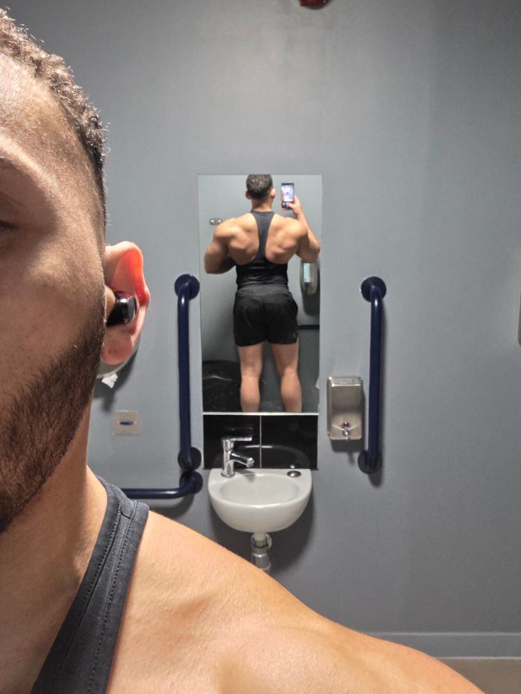 Photo of my back in small mirror is bathroom wearing black vest and shorts