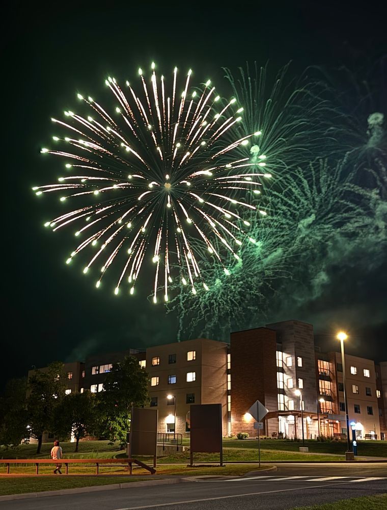 A large green firework explodes in the night sky above a multi-story residence hall on the SUNY Polytechnic Institute campus. The building is warmly lit from within, and a few streetlights and trees frame the scene. A person walks along the sidewalk in the lower left corner, admiring the fireworks.