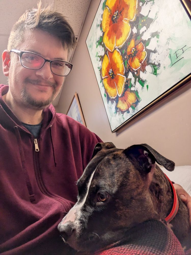 Man in red hoodie, red plaid pajama pants, and black safety glasses looking at camera. A tri-color brindle, boxer-pittbull mix dog, in an orange collar, rests under the man's arm, with his head on the man's knee, looking left. A painting of hibiscus flowers in the background.
