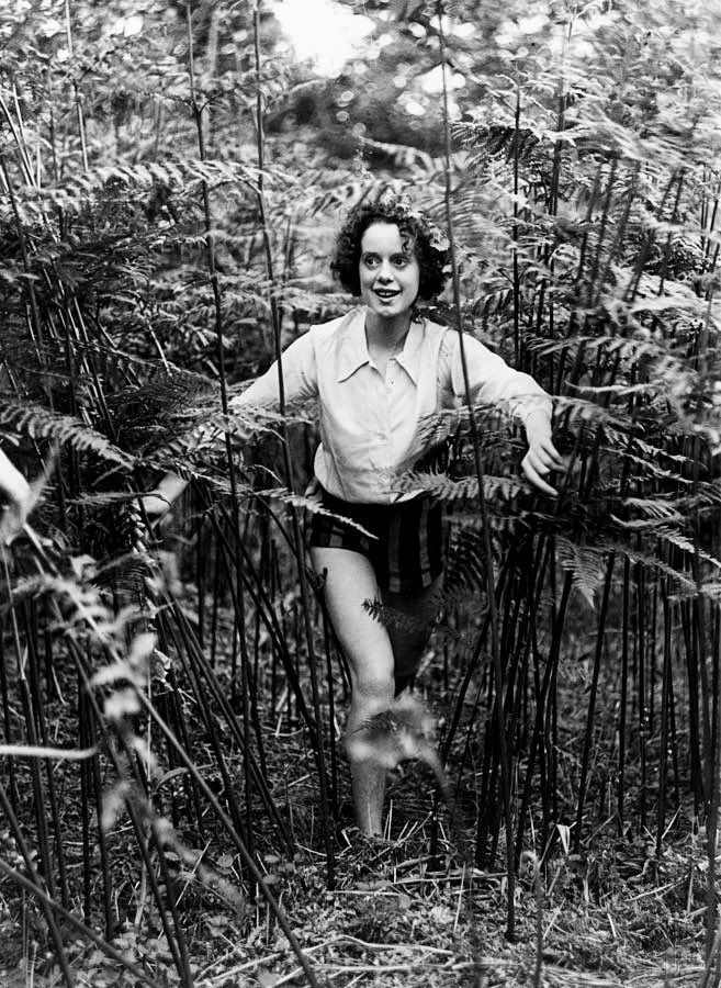 Elsa in a collared white shirt and black shorts, walking through some extremely tall weeds, with a smile on her face 