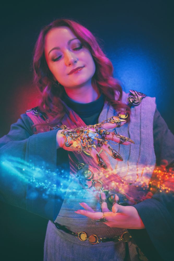 My cosplay of Seldarine drow sorcerer Tav from Baldur’s Gate 3: a woman with long pink hair and purple-shadowed pale skin, in teal, navy, pink, and lavender robes, casting fire and ice magic between her hands while flourishing with a twisted and filigreed golden gauntlet.