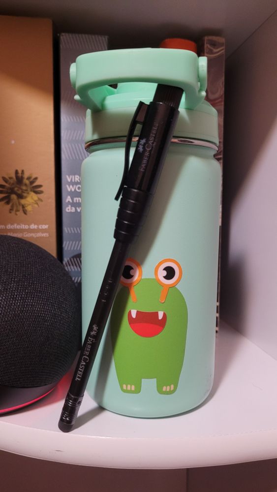 lápis preto faber-castell com borrachinha numa ponta e tampa na outra. na tampa está acoplado um apontador. o lápis está numa estante de livros, apoiada numa garrafinha d'água cor verde claro, na estante há livros e um echo dot.