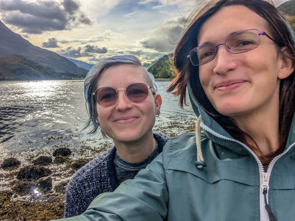 A selfie of a woman with blue hair, left side of head shaved, wearing a warm blue jumper and sun glasses and another woman with normal glasses and brown hair, in a greed raincoat, standing on the bank of a Scottish lake, with water and mountains in the background. Both women are smiling.