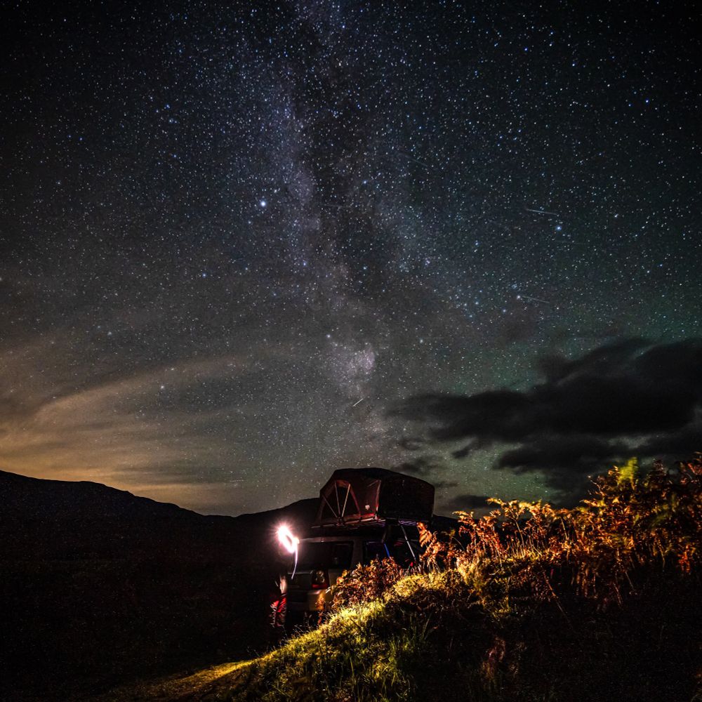 The Milky Way stretched out in the sky above a roof tent, with grasses backlit by torchlight in the foreground 