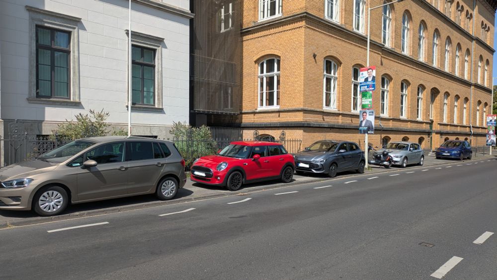 Eine Straße, bei der Gehweg durch mehrere illegal auf dem Gehweg geparkte Autos belegt ist.