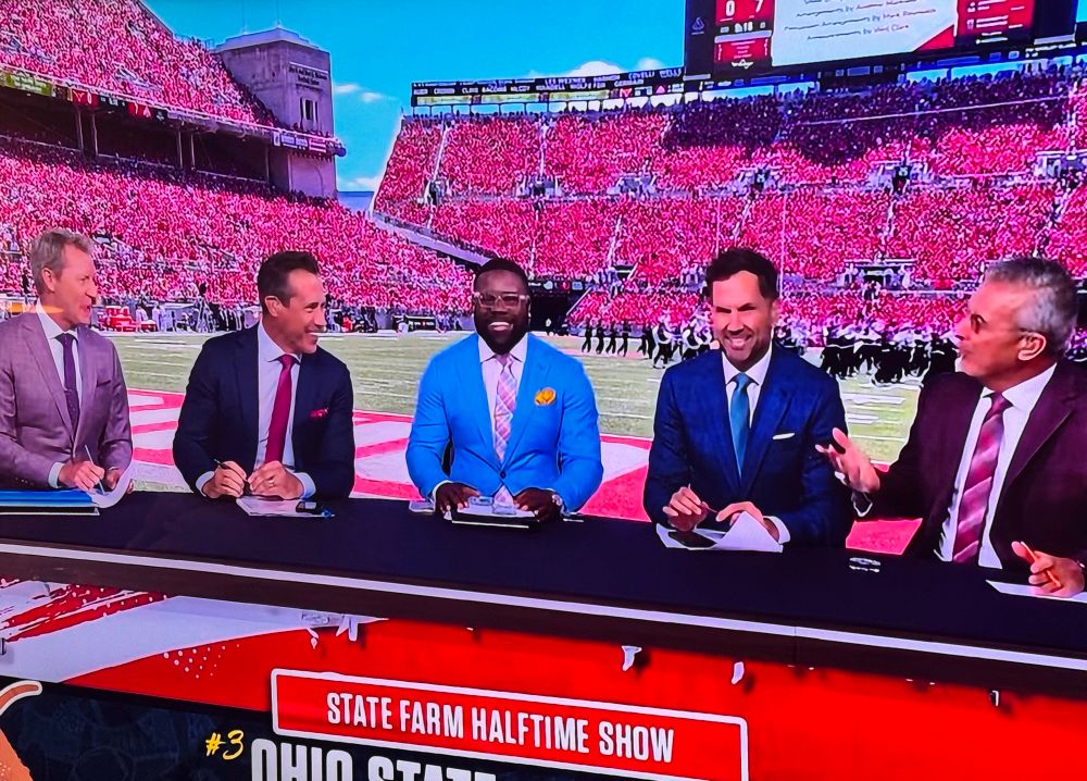 5 men in suits sitting at a table on the field of a college football game. They are wearing too many colors and I already don’t like them. 