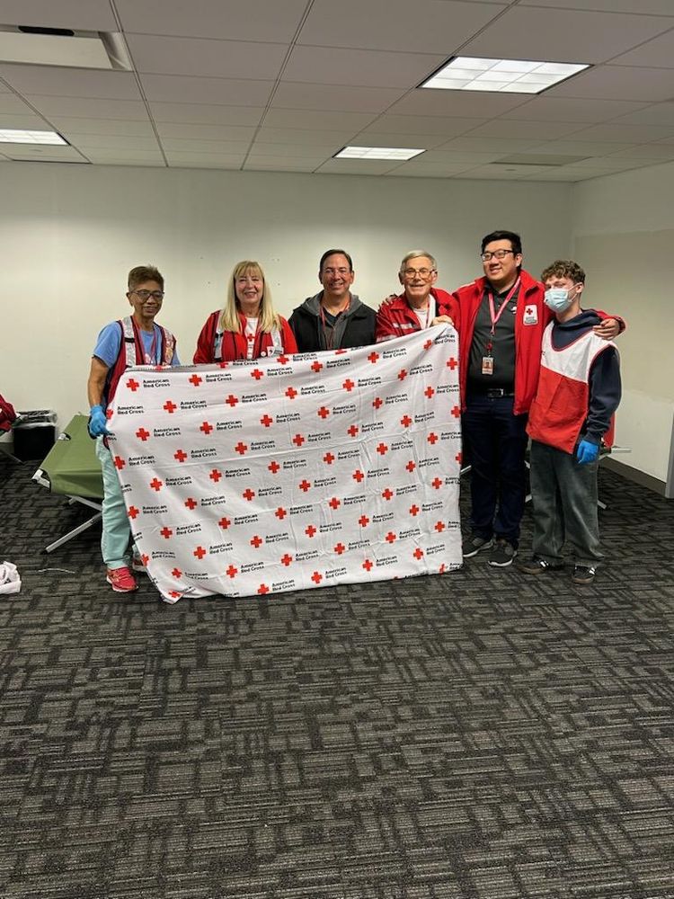 A group of six individuals standing indoors on a dark carpeted floor, holding a large white blanket printed with multiple red crosses and the words “American Red Cross.” Several people are wearing red vests or jackets with Red Cross logos, and some have lanyards and gloves. In the background, there is a folding cot and a plain white wall under a ceiling with fluorescent lights.