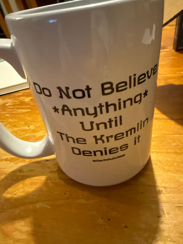 White coffee mug with the text “do not believe *anything* until the kremlin denies it” printed on the side. 