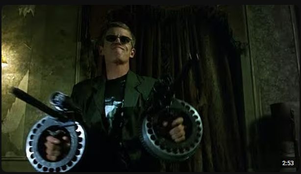 A still from the movie The Matrix of the character Mouse dual wielding custom shotguns. In this still, he looks like Hugh Laurie portraying Dr. House with sunglasses on.