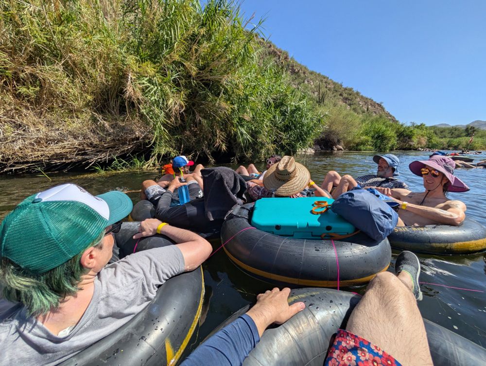 A bunch of GOOD HUMANS in inner tubes floating down a salty river in the desert. There are many colorful hats, jorts, etc.