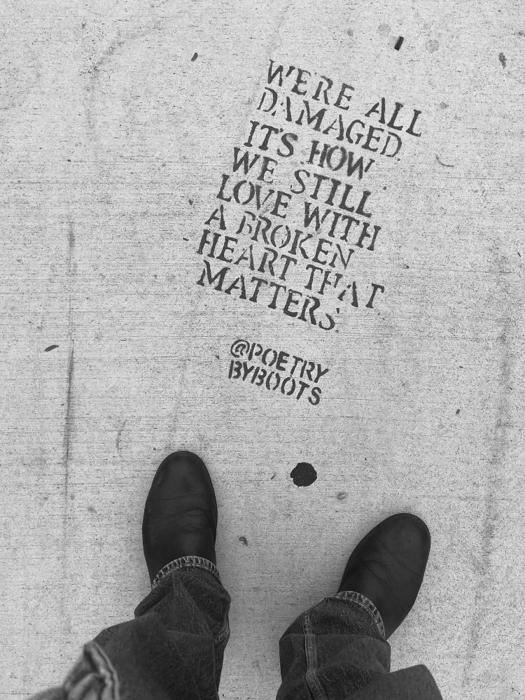 Monochrome. Sidewalk. Boots and jeans in the lower frame. Sidewalk poetry says:

WERE ALL DAMAGED.
IT'S HOW WE STILL LOVE WITH A BROKEN HEART THAI MATTERS
@POETRY
BYBOOTS