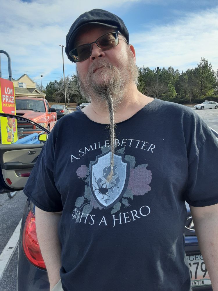 Guy with questionable choice in facial hair wearing a FFXIV shirt that says "A smile better suits a hero"