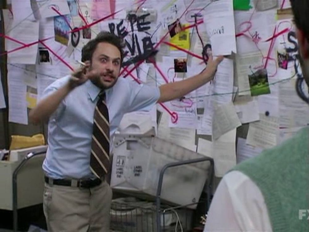 A still from It’s Always Sunny in Philadelphia showing the character Charlie in khakis, a blue button down, and striped tie smoking a cigarette. He’s standing in front of a cork board with papers haphazardly pinned all over it. Circles and red lines suggest connections between these items. Charlie is smoking a cigarette and ranting like a conspiracy theorist. 