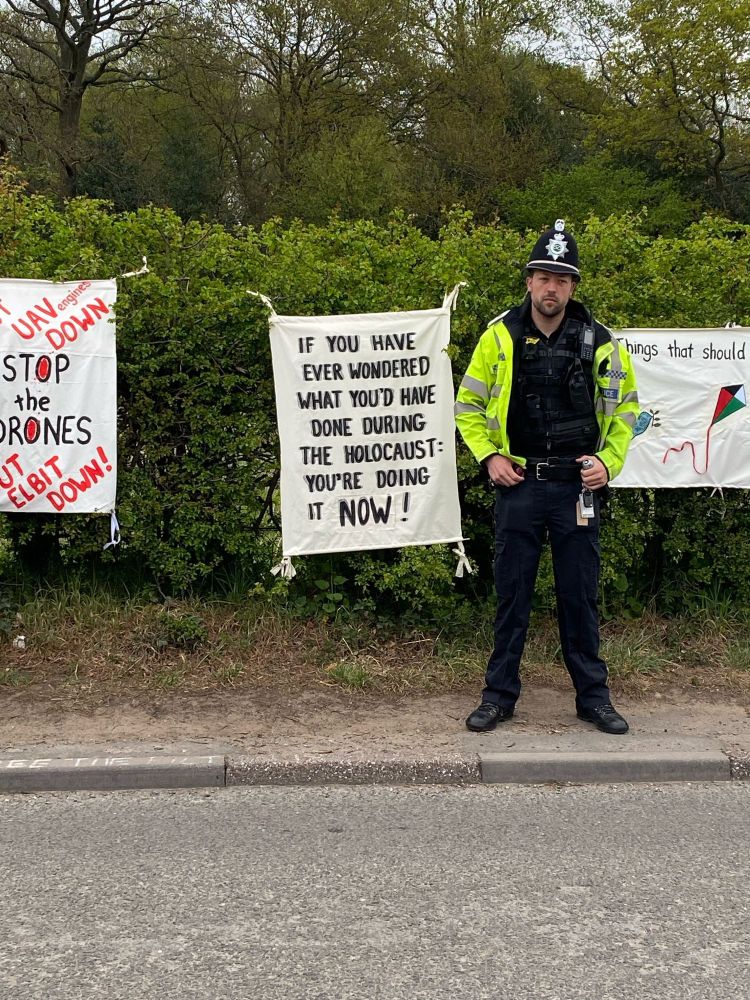 Text on a placard  on a fence:
F YOU HAVE EVER WONDERED WHAT YOU'D HAVE DONE DURING THE HOLOCAUST:
YOU'RE DOING IT NOW!

Next to the sign is a very unfriendly looking police officer from the Staffordshire constabulary.

