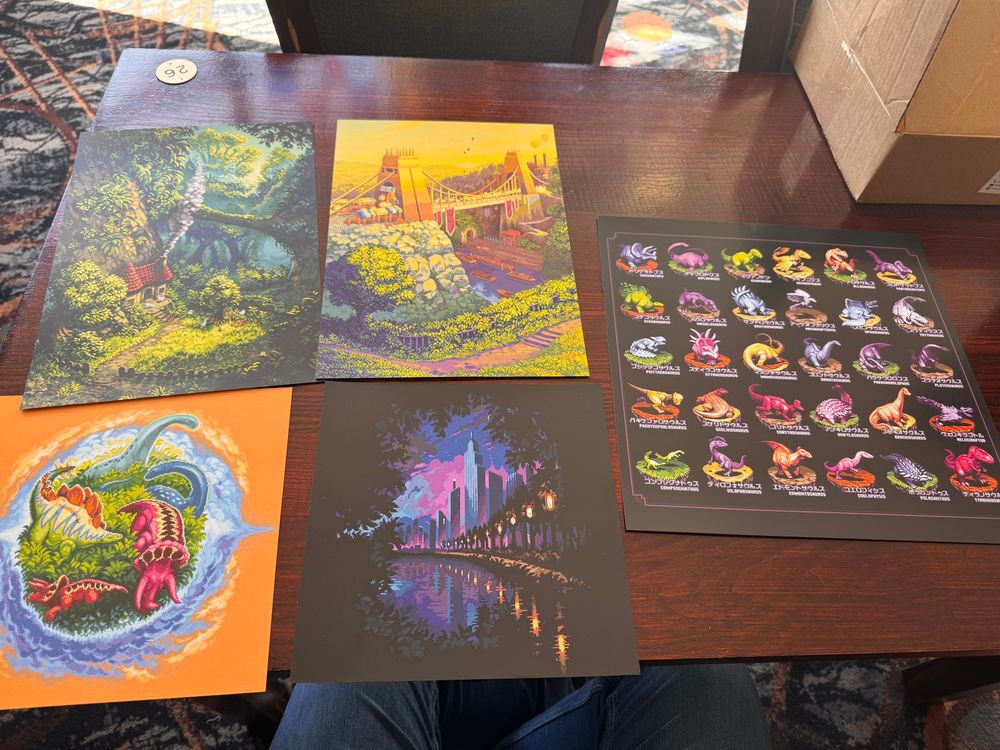 A dark brown tabletop with a selection of pixel art prints on it, including: a forest chine scene with a little house in it, a small forest planet covered with dinosaurs, a cityscape surrounded by a forest vignette, a suspension bridge at evening light, and a chart covered in multiple pixel art dinosaurs. 