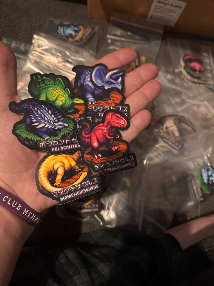 Photograph of my open hand, palm facing up - on the palm rests five dinosaur stickers (Tyrannosaurus, triceratops, Stegosaurus, Mamnchisaurus and Polacanthus). On the floor behind the hand, a big pile of dinosaur sticker sachets are strewn about. 