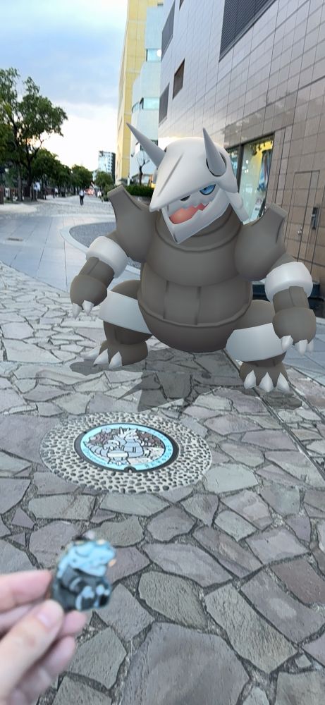The Aggron manhole cover with a (AR) Aggron standing next to it. In the bottom left corner of the picture is my hand holding an Aggron charm based on a design made by @tsurudraws.art 