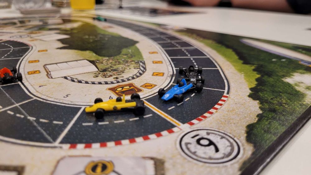 Bild von einem Brettspiel. Man sieht einen Ausschnitt von einem Spielplaan auf dem eine Rennstrecke abgebildet ist und drei kleine Rennwagen, die um eine Kurve fahren.
