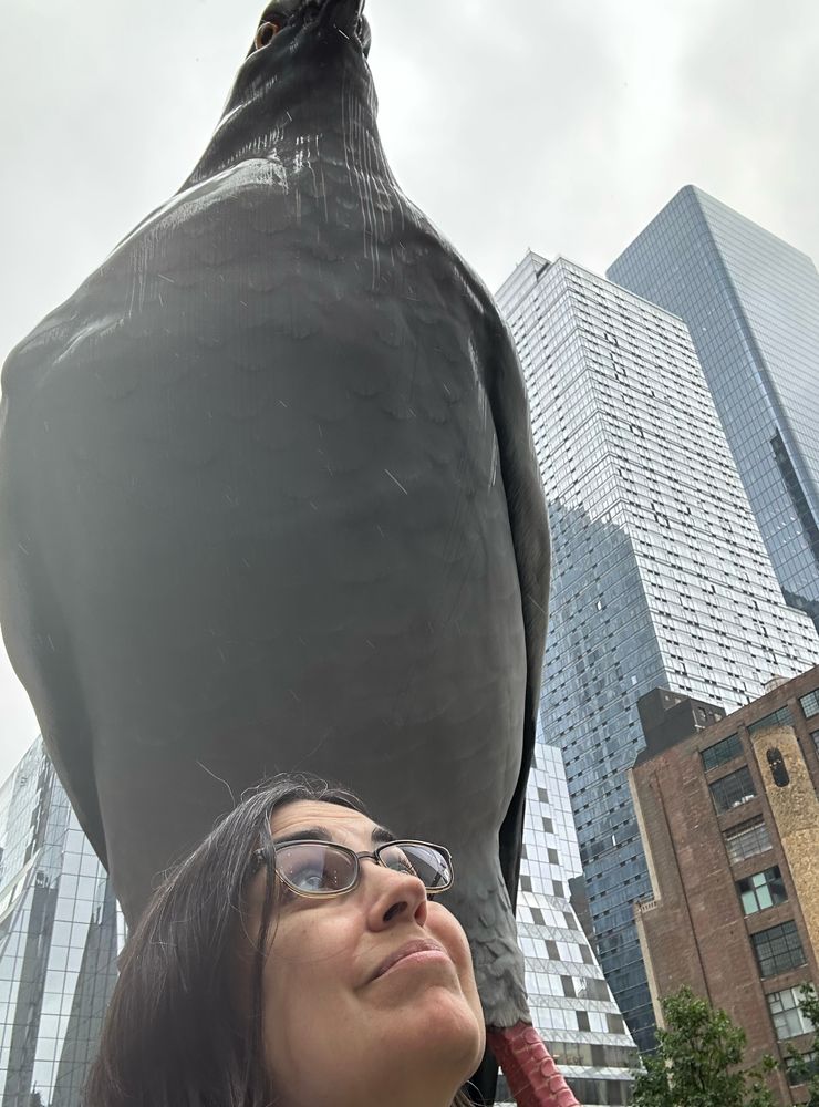 A brown haired woman standing below an enormous statue of a pigeon in NYC.