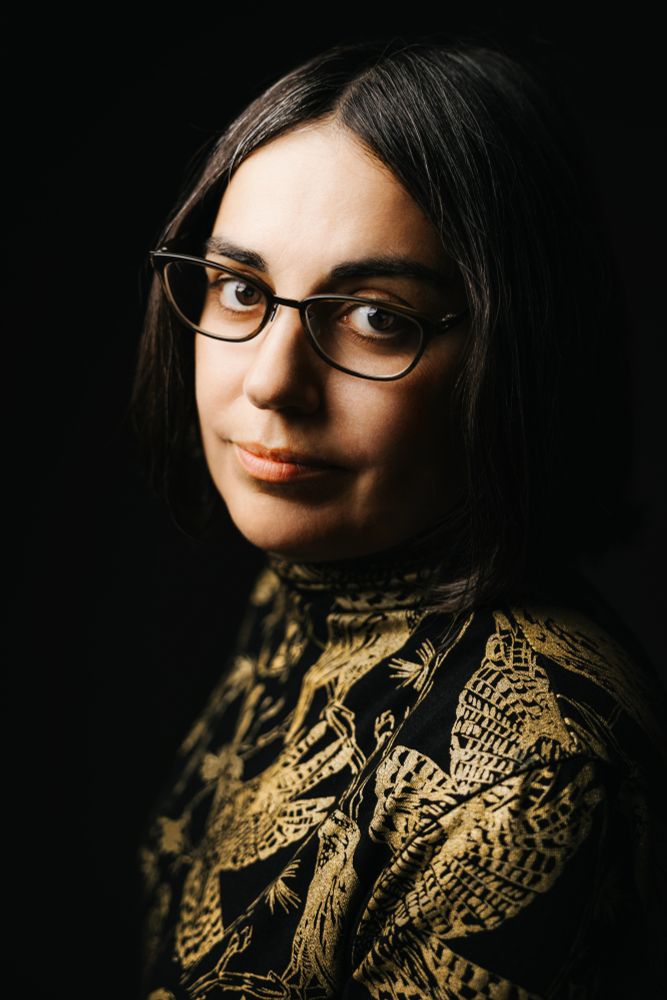 A woman with short brown hair and a black collared shirt with gold birds of prey.