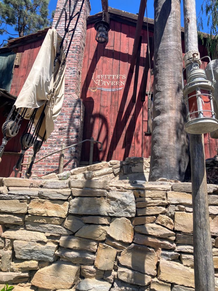 Photo from Tom Sawyer Island at Disneyland. The background is “Lafitte’s Tavern” painted on the side of a red building. In the photo’s foreground is a stone wall, ship’s rigging and a lantern hanging from a post or mast.