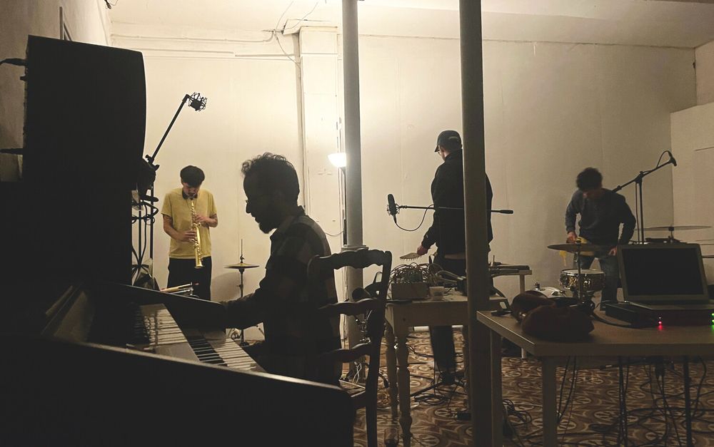 Photo of a Saturday night improv jam in the main studio. The light is low so the four figures appear almost in silhouette. The nearest plays piano, one plays a wind instrument, others percussion.