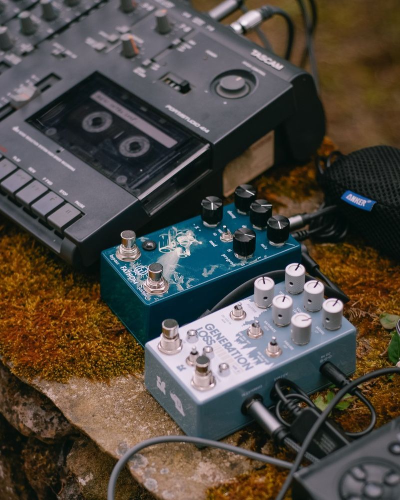 Close-up of Tascam Portastudio 414 and effects pedals on a mossy wall.