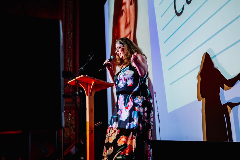 Me performing at the Mortified Chicago storytelling series. A white woman with red hair and glasses, wearing a floral dress, stands in front of a movie screen speaking into a microphone. 