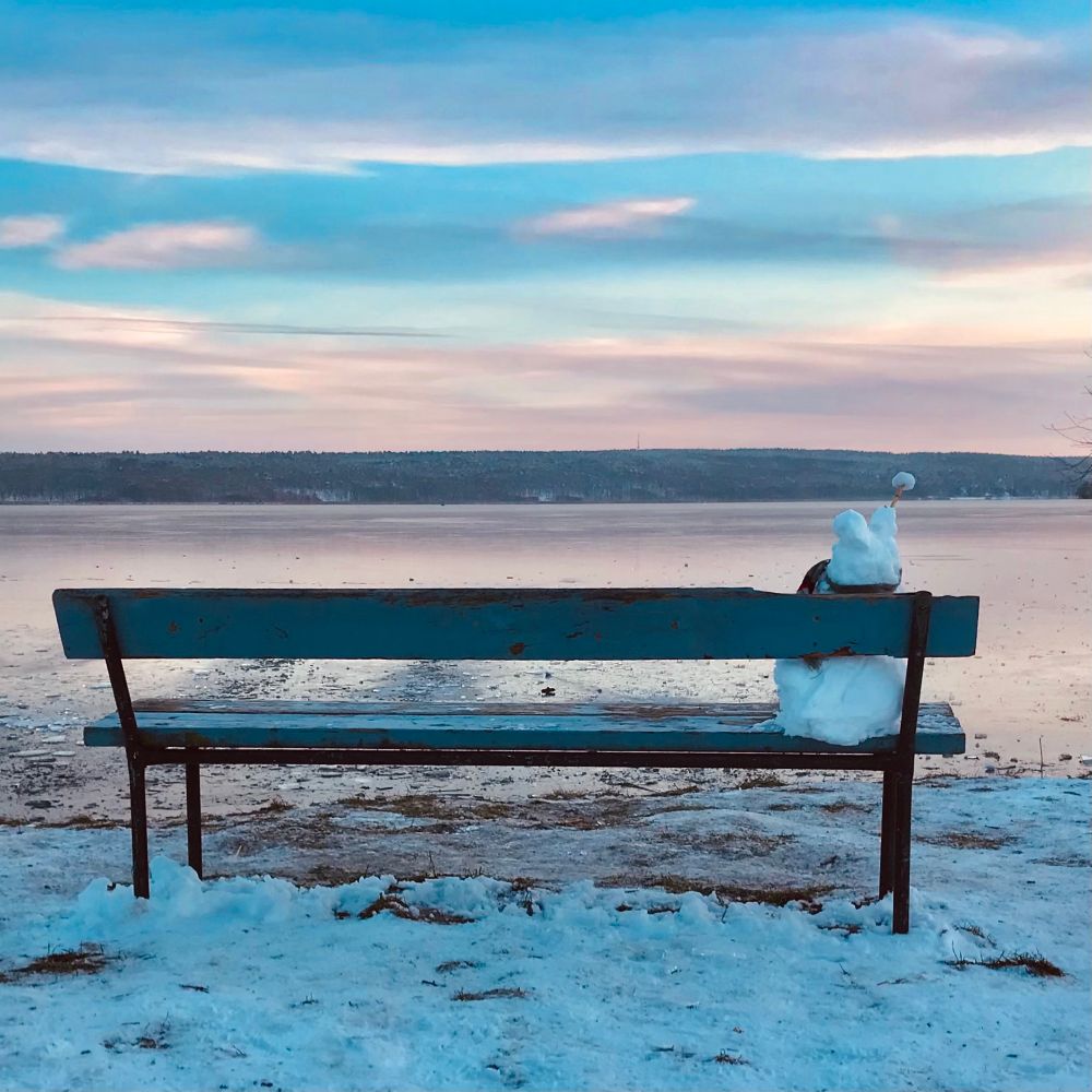 Auf einer Bank sitzt ein kleiner Schneemann und blickt auf einen zugefrorenen See. Der Himmel ist blau-rosa gefärbt. Im Vordergrund liegt Schnee 
