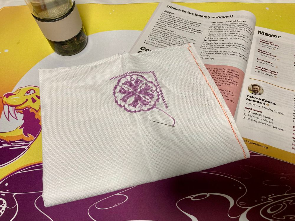 more of the Quilt Sample cross-stitch has been completed. It sits on top of the NYC 2025 voter guide, both of those on top of a orange/purple Frame Fatales desk mat; an infusion tumbler of cherry green tea sits off to the top left