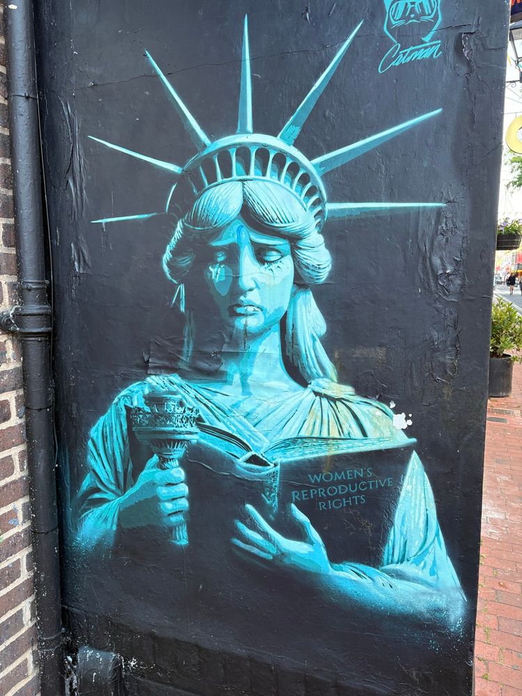 A graffiti of the Statue of Liberty looking sad and looking down into a book. The title of the book reads “Women’s reproductive rights”. Found in The Lanes in Brighton.