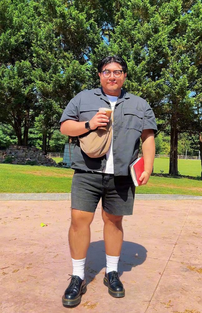 Ed wearing a black button down with a white shirt underneath, black shorts and white socks and black Dr.Martens. He’s holding a book and coffee.