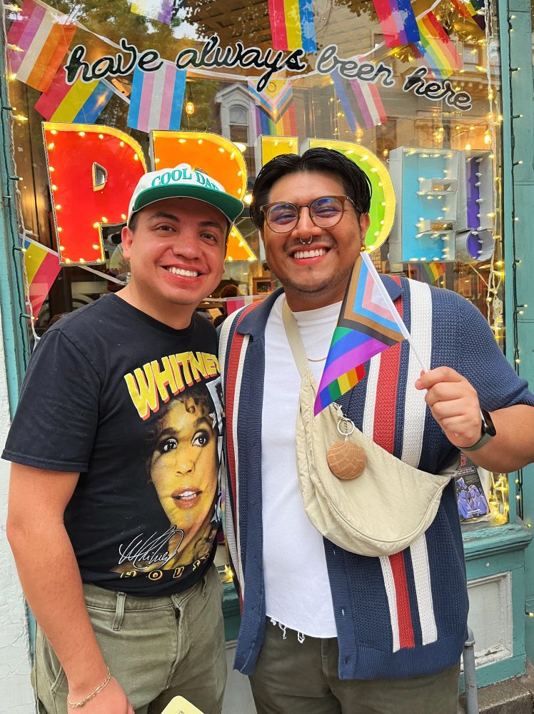 Ed and his bf at a pride parade. Ed is holding a pride flag and they’re standing in front of a Pride-themed window display.
