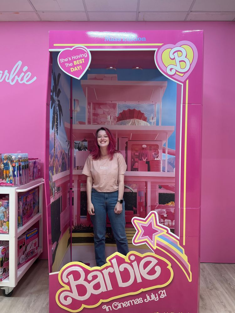 Jodie, a girl with pink hair wearing a pink T-shirt and blue jeans is smiling inside a life-sized pink Barbie box.