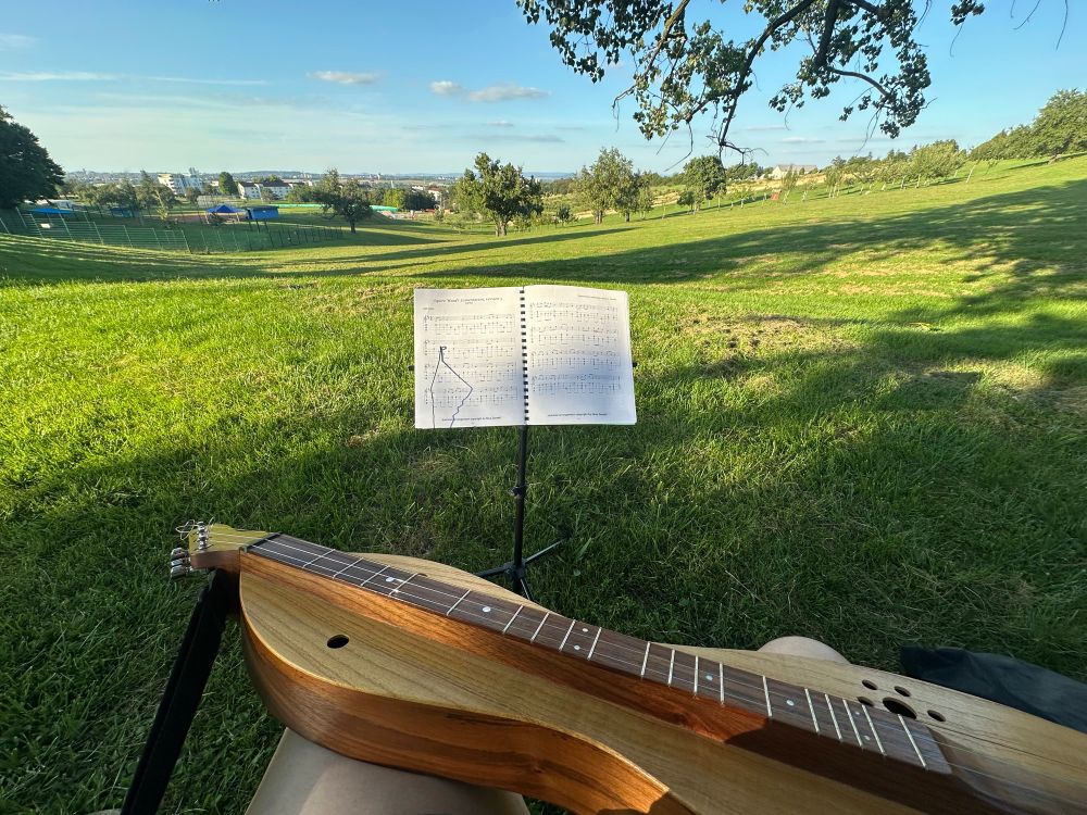 A countryside vista with an orchard and clouds in a mid-August day. In the foreground is a metal music stand with an open music book. A mountain dulcimer emerges in the immediate foreground. The grass is dappled with shadows from large trees and clouds. 
