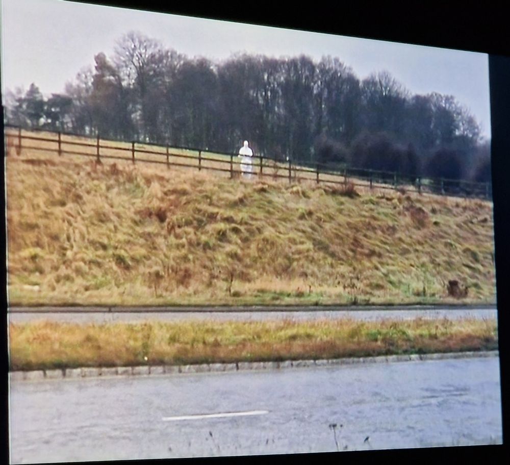 A mysterious white figure stands in the distance on the other side of a bypass.