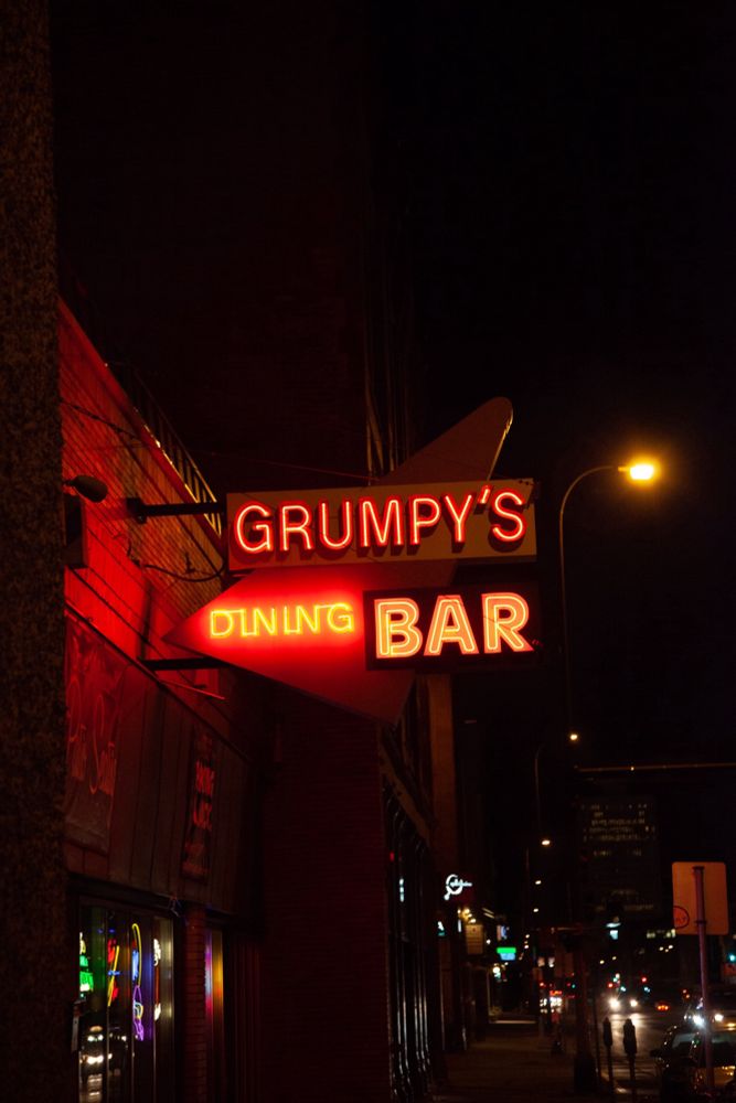 A photo of a space age neon sign reading “Grumpy’s Dining Bar” - taken at night with a storefront filled with brewery signs slightly visible below. 