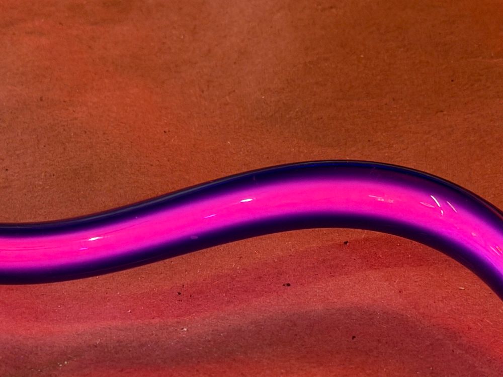 A wavy neon sign tube on a workbench. The tube is a deep violet blue and dark but the plasma center of the tube is glowing a deep magenta color. 