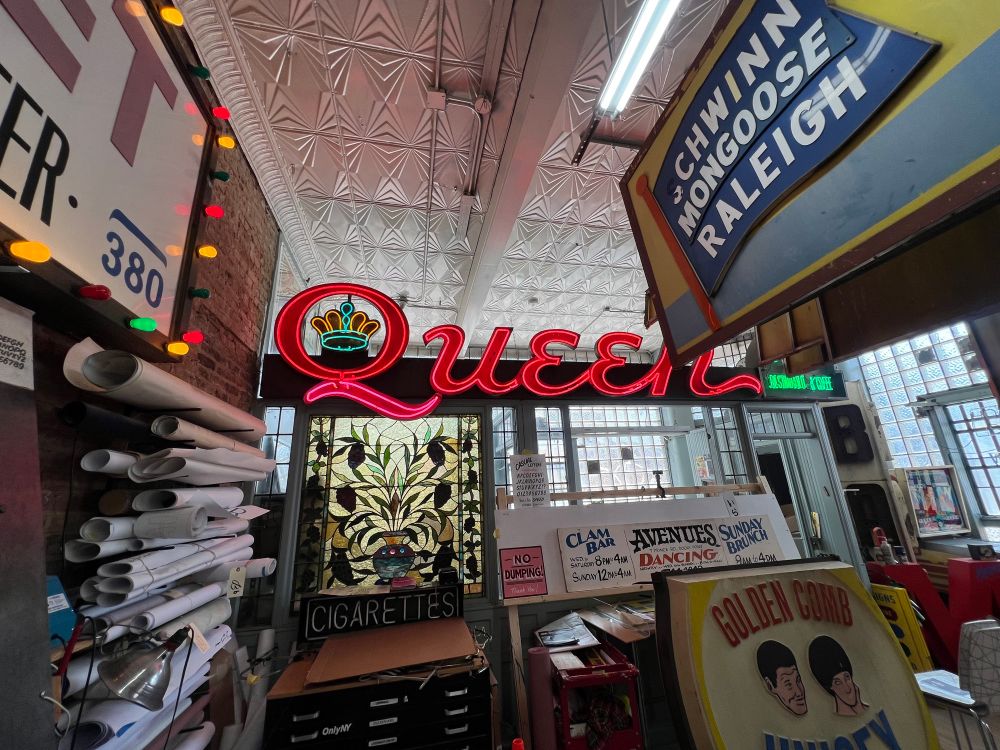 A pink neon “Queen” sign burring behind many old panel storefront signs hanging from the ceiling. The word is in an all capital script and the Q has a little crown in the negative space.