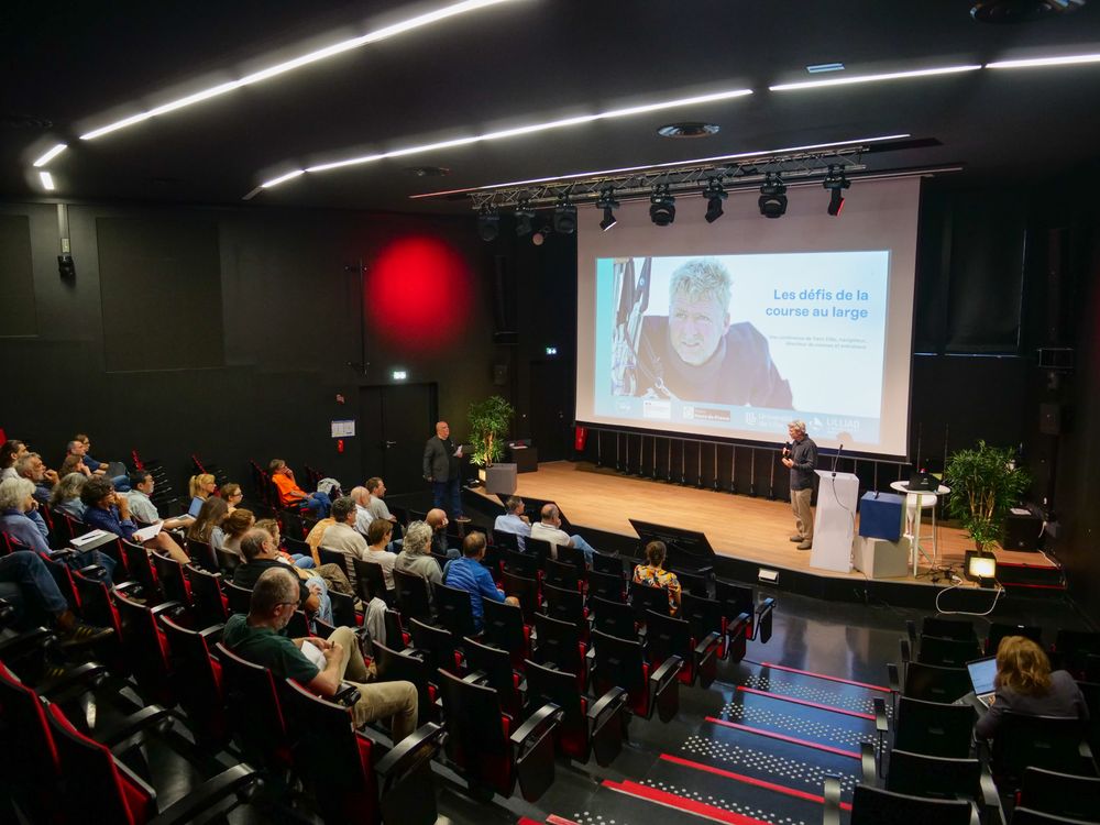 Un amphithéâtre avec des personnes assises. Le conférencier Yann Eliès est sur scène pour faire sa présentation. À l'écran, une photo du conférencier sur un bateau et le titre de la conférence "Les défis de la course au large".
