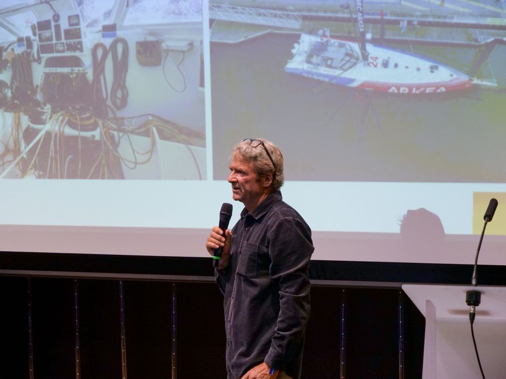 Le conférencier Yann Eliès est sur scène pour faire sa présentation. Deux photos sont affichées sur l'écran derrière lui. À gauche, une vue intérieure d'un bateau : un tableau de bord avec de nombreux câbles et instruments électroniques. À droite, une vue extérieure d'un bateau à voile amarré. 