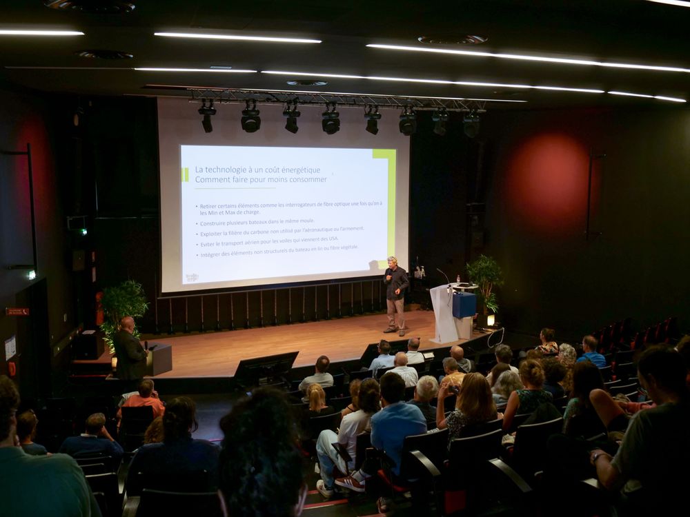 Un amphithéâtre avec des personnes assises. Le conférencier Yann Eliès est sur scène pour faire sa présentation. Sur l'écran situé derrière lui, une diapositive intitulée "La technologie a un coût énergétique. Comment faire pour moins consommer ?" est projetée. 