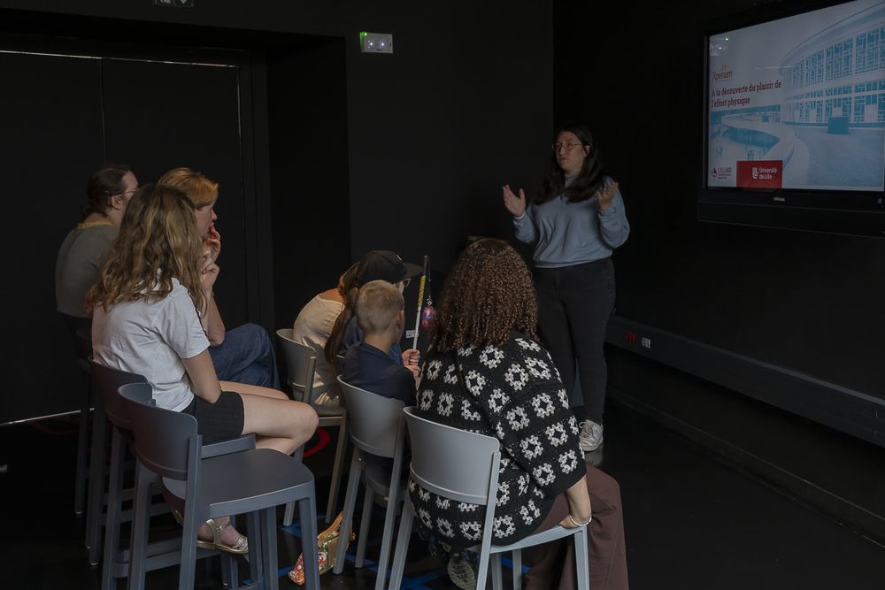 Un groupe d'adolescents est assis sur des chaises. Ils écoutent une animatrice du stand "à la découverte du plaisir de l'effort physique".