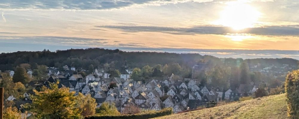 Das Bild zeigt eine deutsche Kleinstadt. Die Häuser sind traditionelle Fachwerkhäuser mit dunklen Balken und weißen Wänden. Im Hintergrund erstreckt sich ein bewaldeter Hügel, der die Szene einrahmt. Der Himmel ist hell und wird von der Sonne beleuchtet, die in der Ferne durch die Wolken scheint. Das Licht verleiht dem Bild eine warme und einladende Atmosphäre.