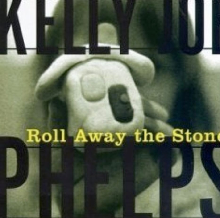 Das Bild zeigt das Cover des Albums „Roll Away the Stone“ von Kelly Joe Phelps. Im Vordergrund ist ein aus Ton geformtes Haus zu sehen, das den Großteil des Covers ausfüllt. Das Haus verdeckt einen menschlichen Kopf, der dahinter verschwommen zu erkennen ist. Der Name des Künstlers und der Albumtitel sind in großen, fettgedruckten Buchstaben darüber und darunter platziert. Das Gesamtbild ist in Sepia-Tönen gehalten und wirkt etwas düster und geheimnisvoll.