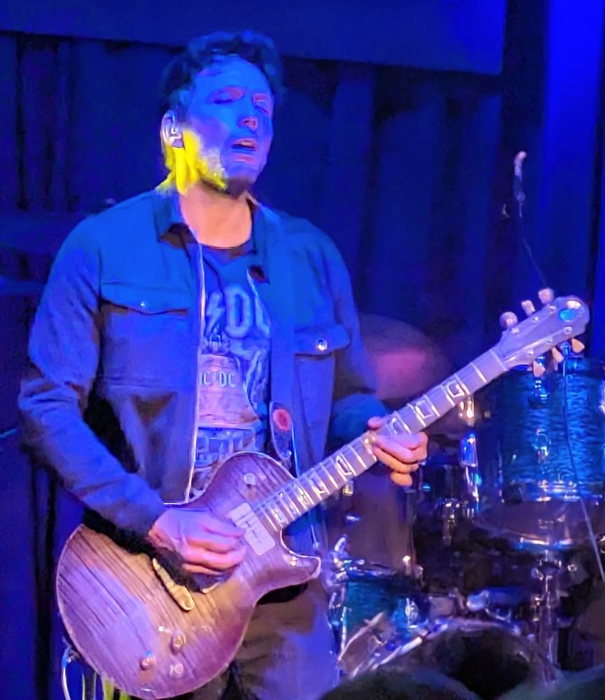 Das Bild zeigt Aynsley Lister,  der eine Patrick James Eggle Macon Single Cut spielt. Er trägt ein T-Shirt mit einem AC/DC-Logo und eine blaue Jacke. Im Hintergrund sieht man einen Teil eines Schlagzeugs. Die Beleuchtung ist in Blau- und Gelbtönen gehalten.