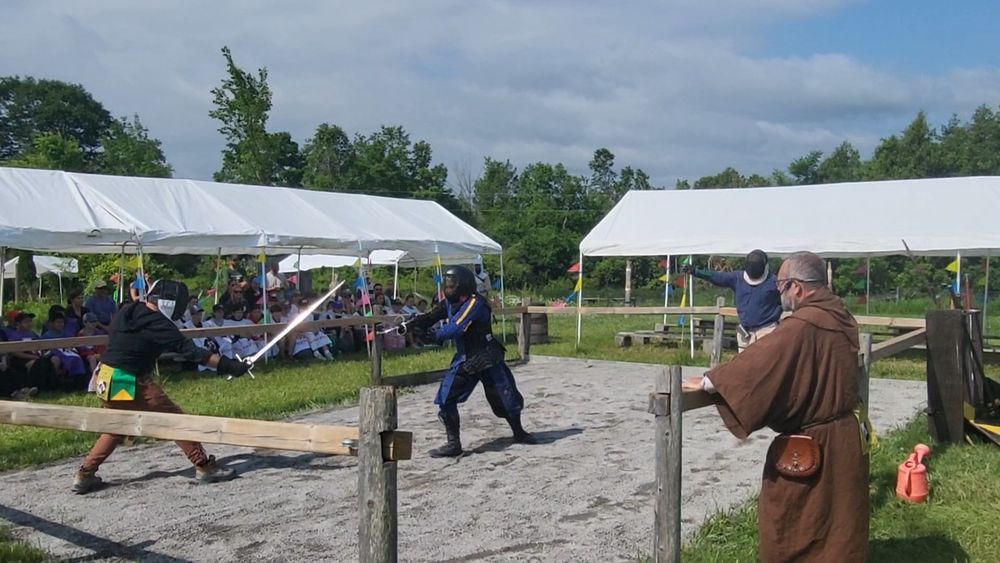 The woman in the blue outfit is now wearing a helmet. She is between two swordsmen, defending herself with her greatsword.  An audience is watching  the demonstration.