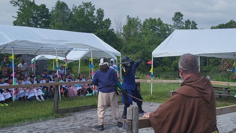The woman in the blue outfit has her greatsword in the fencing mask of an opponent, who is wielding a spear. An audience is watching this demonstration.